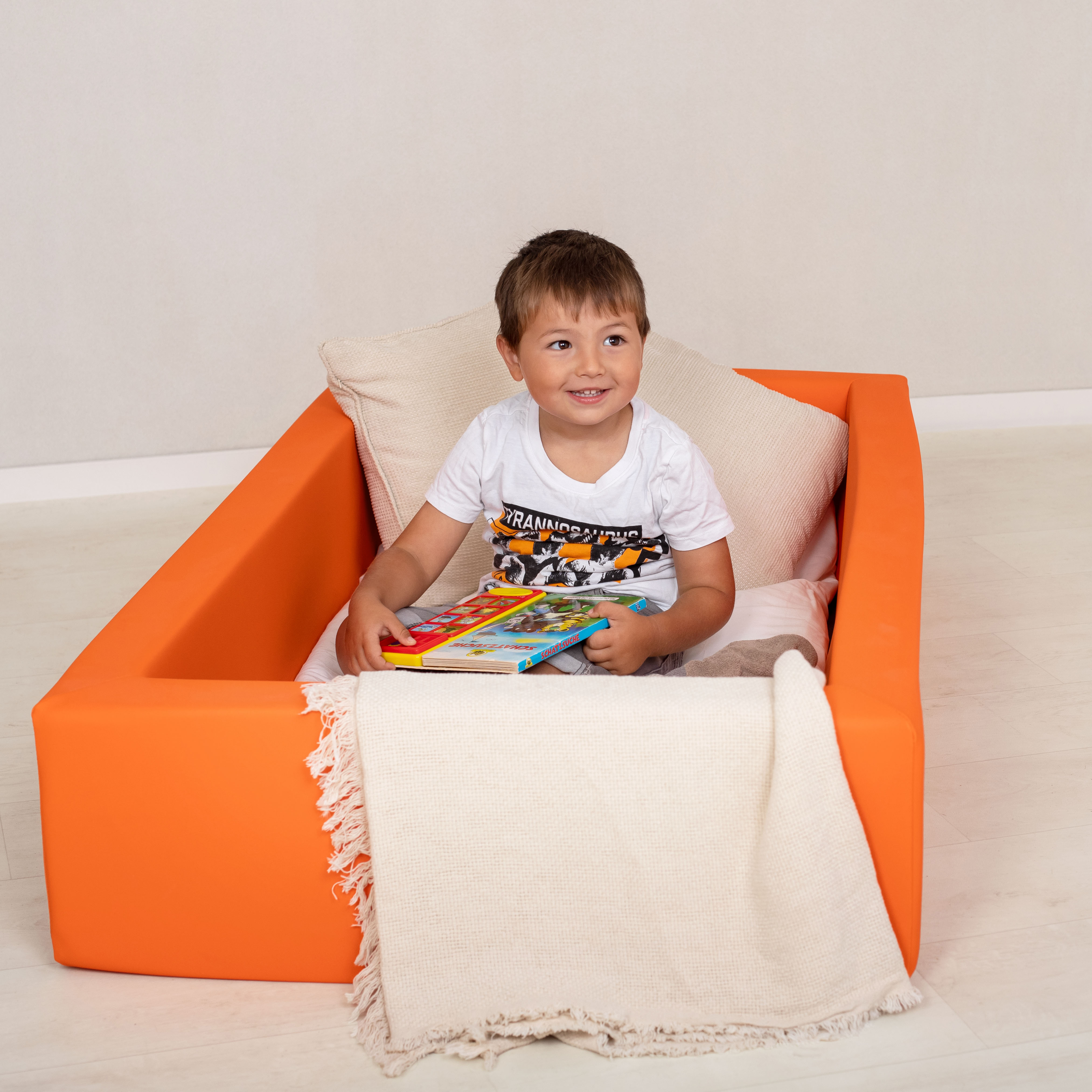 Kleiner Junge sitzt in einem orangen Kinderbett aus Vollschaum  liest ein Kinderbuch.